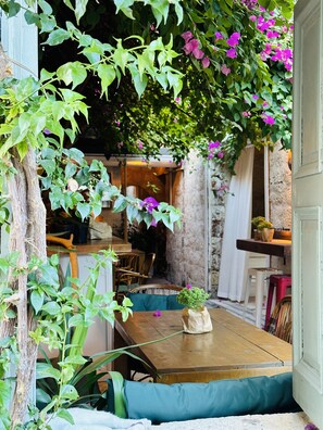 An outdoor seating area with a wooden table and blue cushions, surrounded by lush greenery and purple flowers. - Alaca Butik Otel (Cesme)