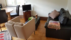 Ett vardagsrum med en svart soffa, en beige fåtölj, ett träbord, en tv-apparat på ett träställ och en liten trumma.