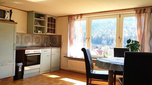 Ett kök med mikrovågsugn, ugn och skåp. Det finns ett fönster med gardiner och utsikt över träd.
