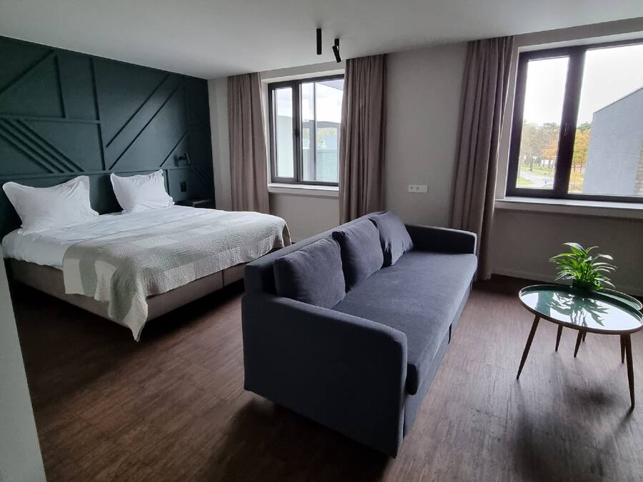Een moderne kamer met een bed, een bank, een kleine tafel met een plantje en grote ramen.