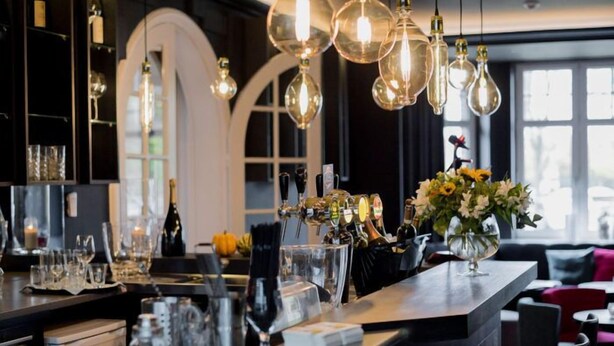 A bar area with a counter, wine glasses, bottles, and a flower arrangement. - Hotel Porta Cite (Maasmechelen)