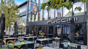 An outdoor seating area with tables and chairs, a storefront with 'PUCCINI' signage, and a tree in the foreground. - Hotel Porta Cite (Maasmechelen)