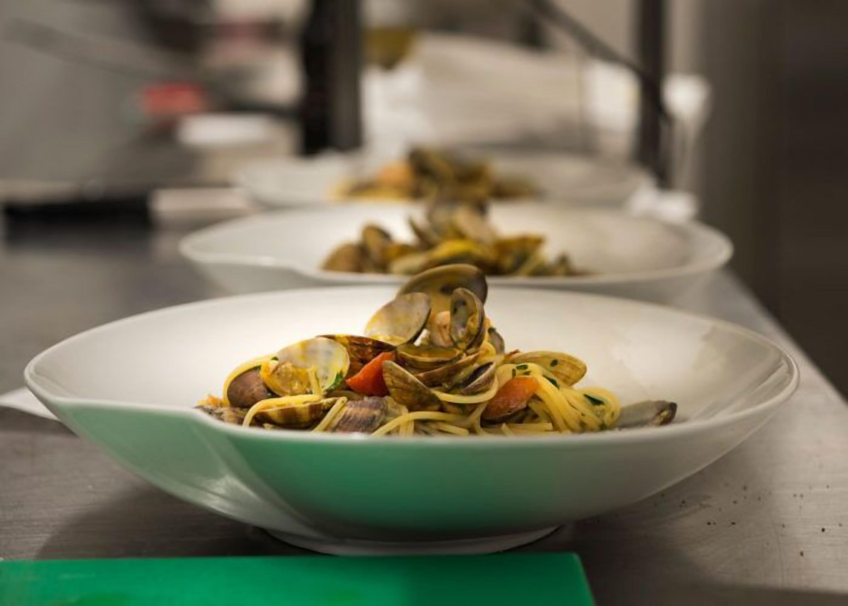 Una fila de platos blancos con pasta de mariscos sobre una encimera de acero inoxidable.