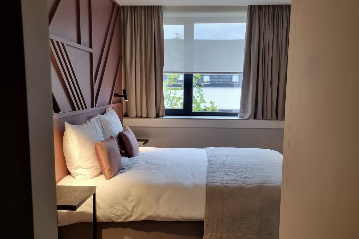 A neatly made bed with a headboard, a bedside table, and a window with curtains.