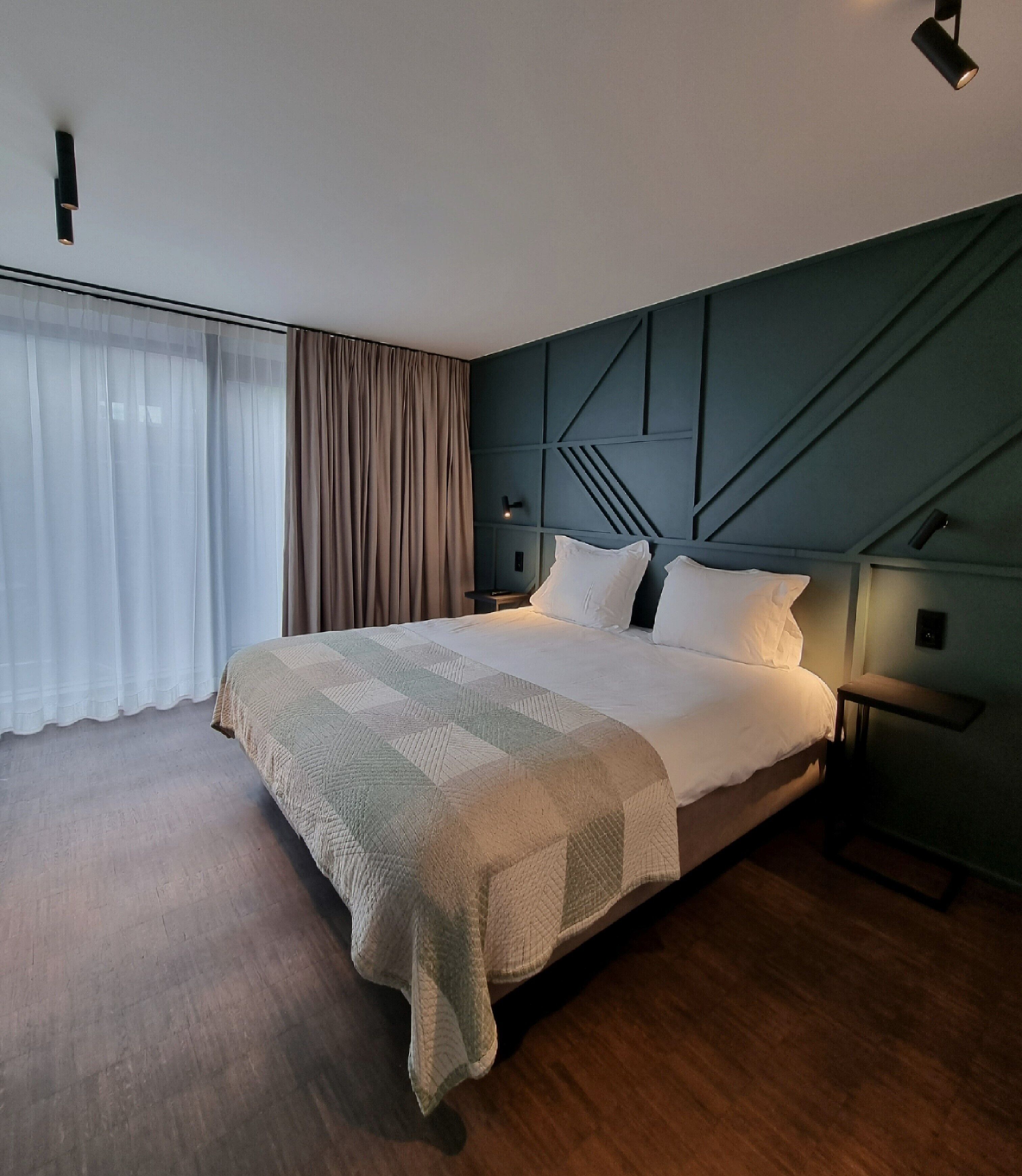 Ein modernes Hotelzimmer mit einem großen Bett, einem Nachttisch, einer Wandleuchte und transparenten Vorhängen.