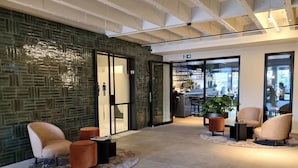 A modern lobby with a green mosaic wall, seating area with armchairs and ottomans, and a bar area visible through glass doors. - Hotel Porta Cite (Maasmechelen)
