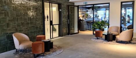 A modern lobby with a green mosaic wall, seating area with armchairs and ottomans, and a bar area visible through glass doors.