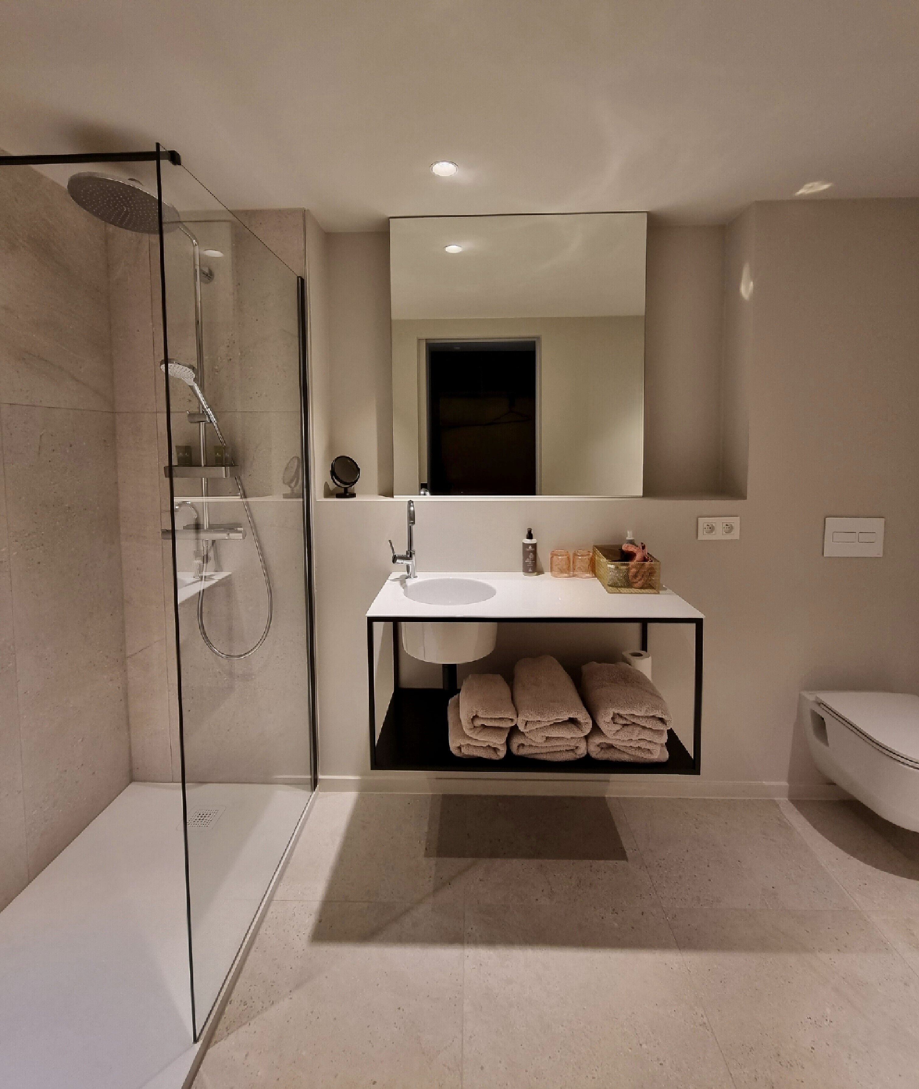 A modern bathroom with a glass-enclosed shower, a white sink, and a mirror above it. There are towels stacked on a shelf below the mirror.