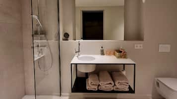 Une salle de bain moderne avec une douche à parois de verre, un lavabo blanc et un miroir au-dessus. Des serviettes sont pliées et rangées sur une étagère située sous le miroir.