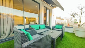 A modern outdoor seating area with wicker furniture and green cushions, large windows, and a view of the cityscape.