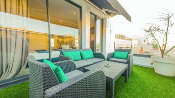 Un área de estar exterior moderna con mobiliario de mimbre y cojines verdes, amplias ventanas y vistas panorámicas de la ciudad.