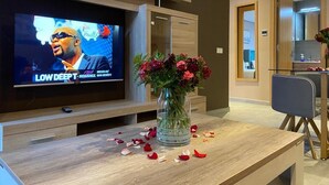 A modern living room with a flat-screen TV, a wooden coffee table with a vase of flowers and rose petals, and a dining area with a chair and table. - Atlas-Résidence By Rent-Inn (Rabat)