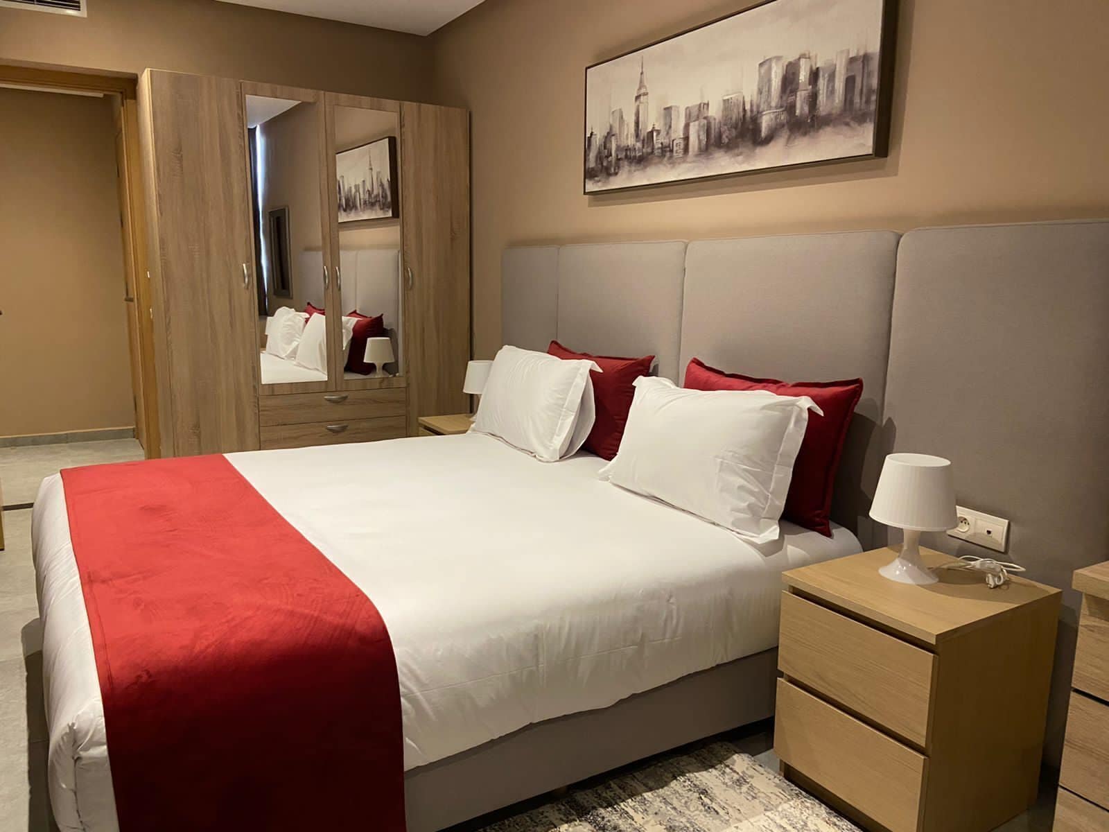 Ein Hotelzimmer mit einem großen Bett, einem Nachttisch, einem Spiegel und einem Stadtbild an der Wand.