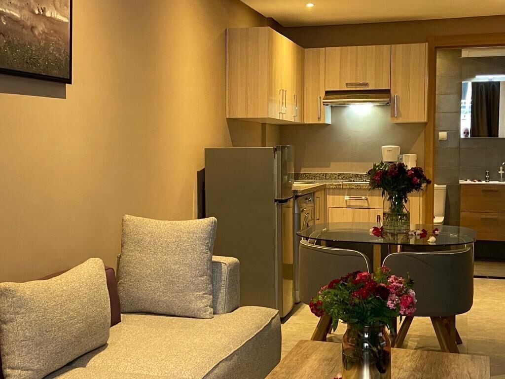 Een knus woongedeelte met een bank, een eettafel met stoelen, een keuken met kastjes en een koelkast, en een kleine decoratieve vaas met bloemen.