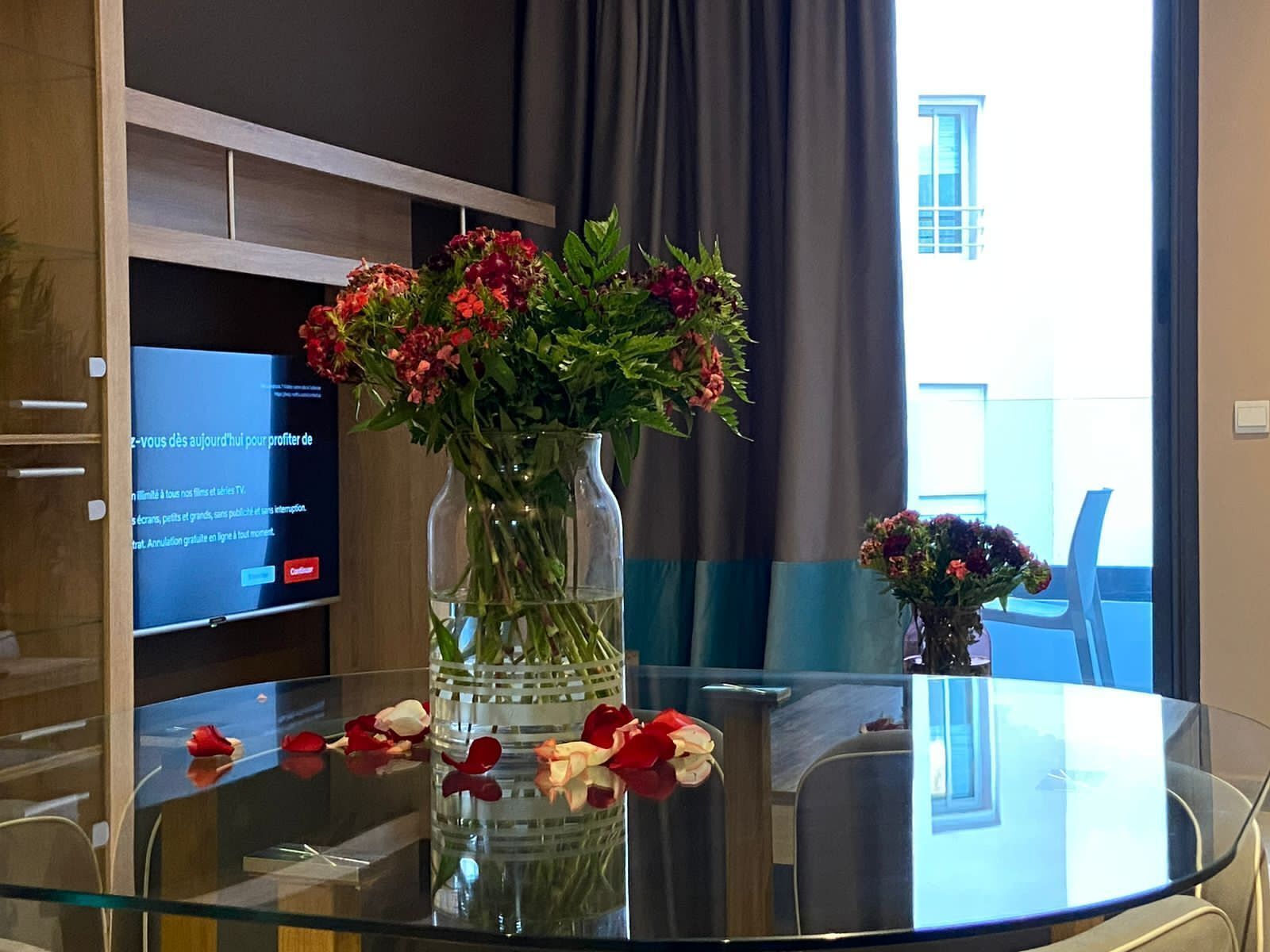 Ein Tisch mit Glasplatte, eine Blumenvase und Rosenblüten, ein Fernsehbildschirm und ein Blick auf ein Gebäude durch ein Fenster.