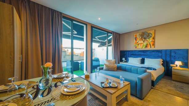 A hotel room with a bed, a sofa, a dining table set with plates and glasses, and a painting on the wall. - Atlas-Résidence By Rent-Inn (Rabat)