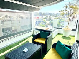 Un balcón con mobiliario de mimbre, una planta en maceta y vista panorámica de la ciudad.