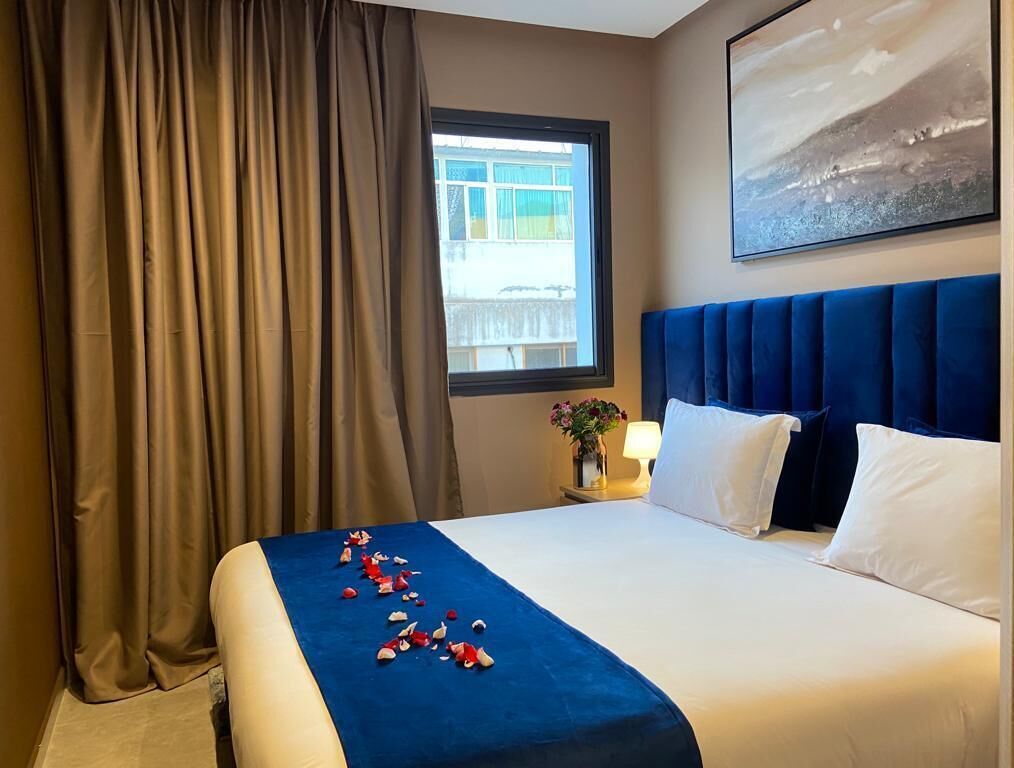 A hotel room with a large bed, a window with curtains, a blue headboard, and a painting on the wall.
