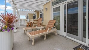Un espace extérieur abrité, doté de chaises longues et d’une table, d’une plante en pot aux fleurs violettes et offrant une vue sur la mer à travers une porte vitrée.