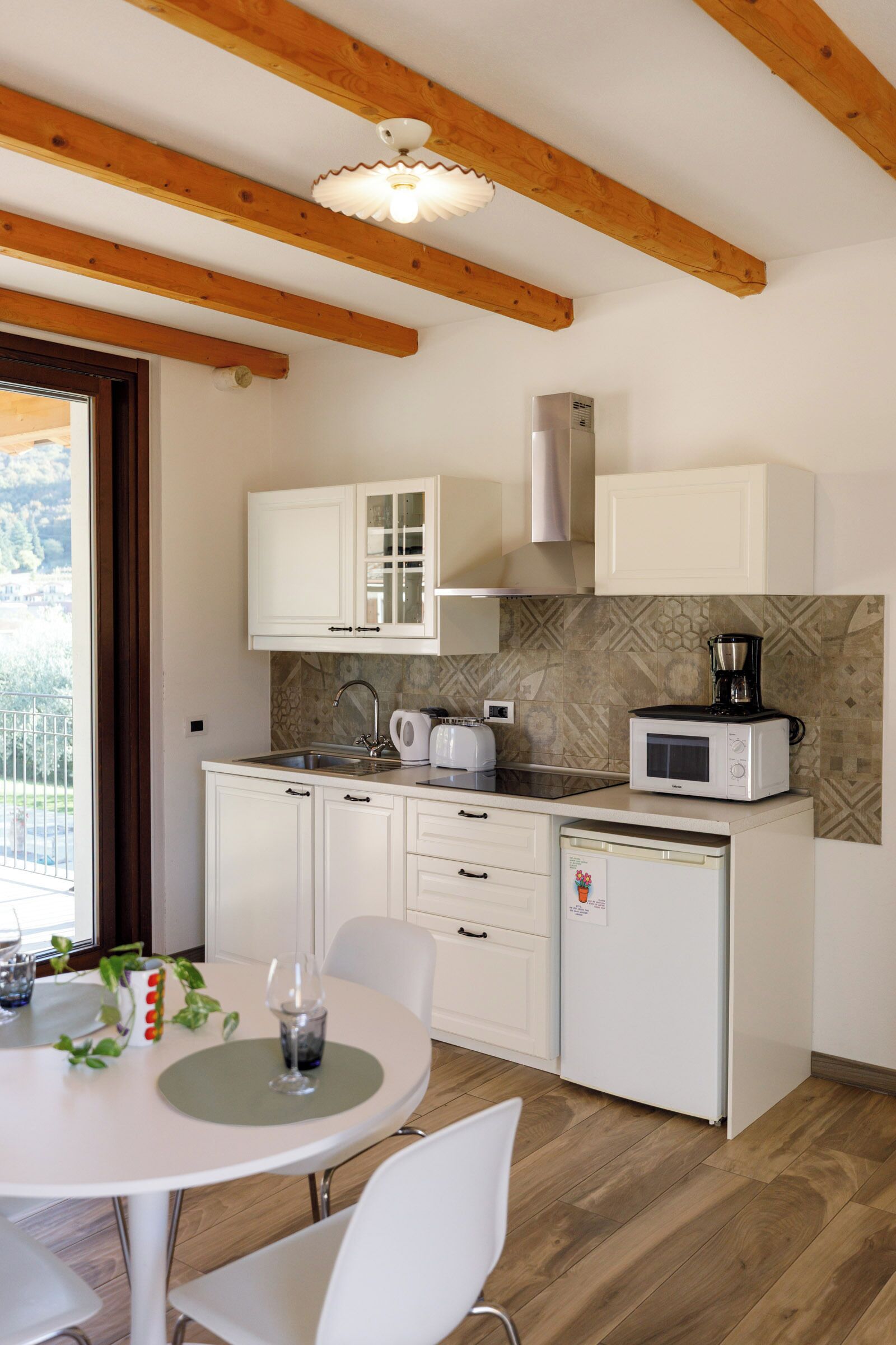 A compact kitchen with white cabinets, a microwave, a coffee maker, and a sink. There is a dining table with chairs and a window with a view of greenery.