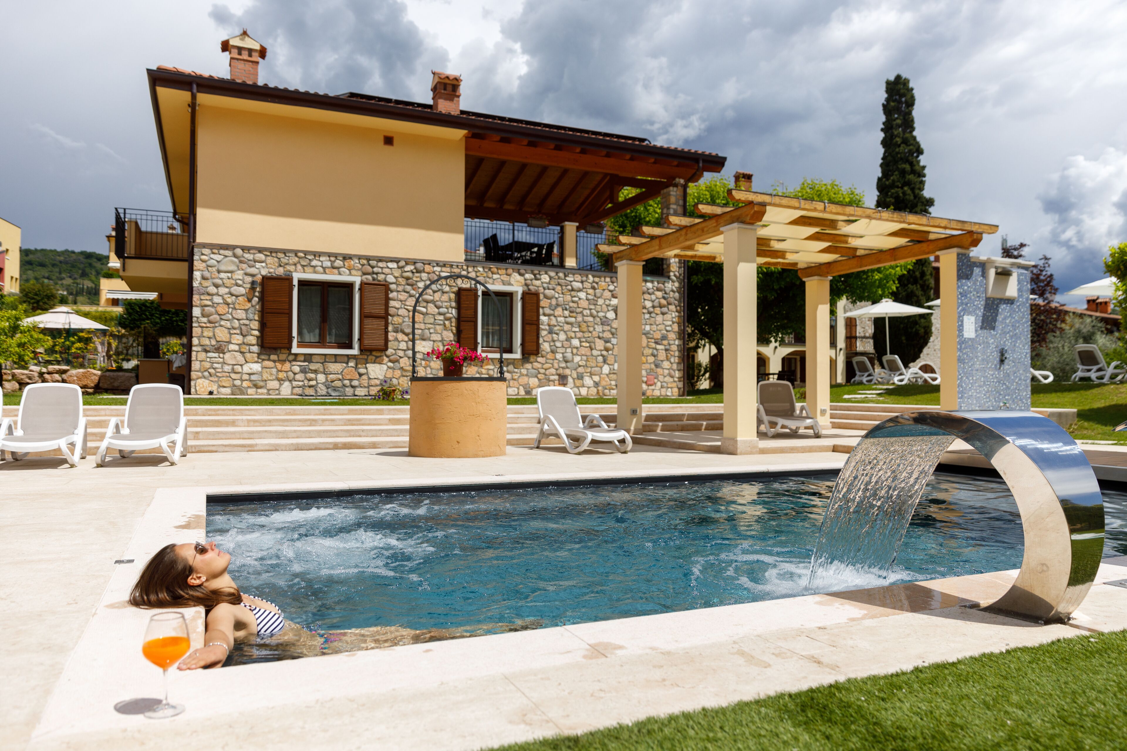 A swimming pool with a water feature, surrounded by lounge chairs and a pergola.