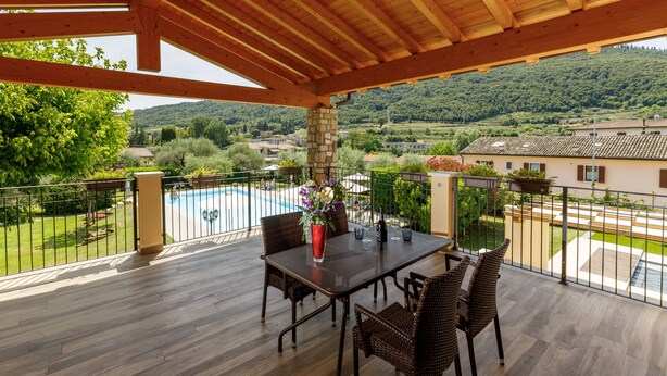 Un espace extérieur abrité, doté d’un plafond en bois, d’une table avec des chaises, et offrant une vue sur une piscine et un espace verdoyant.