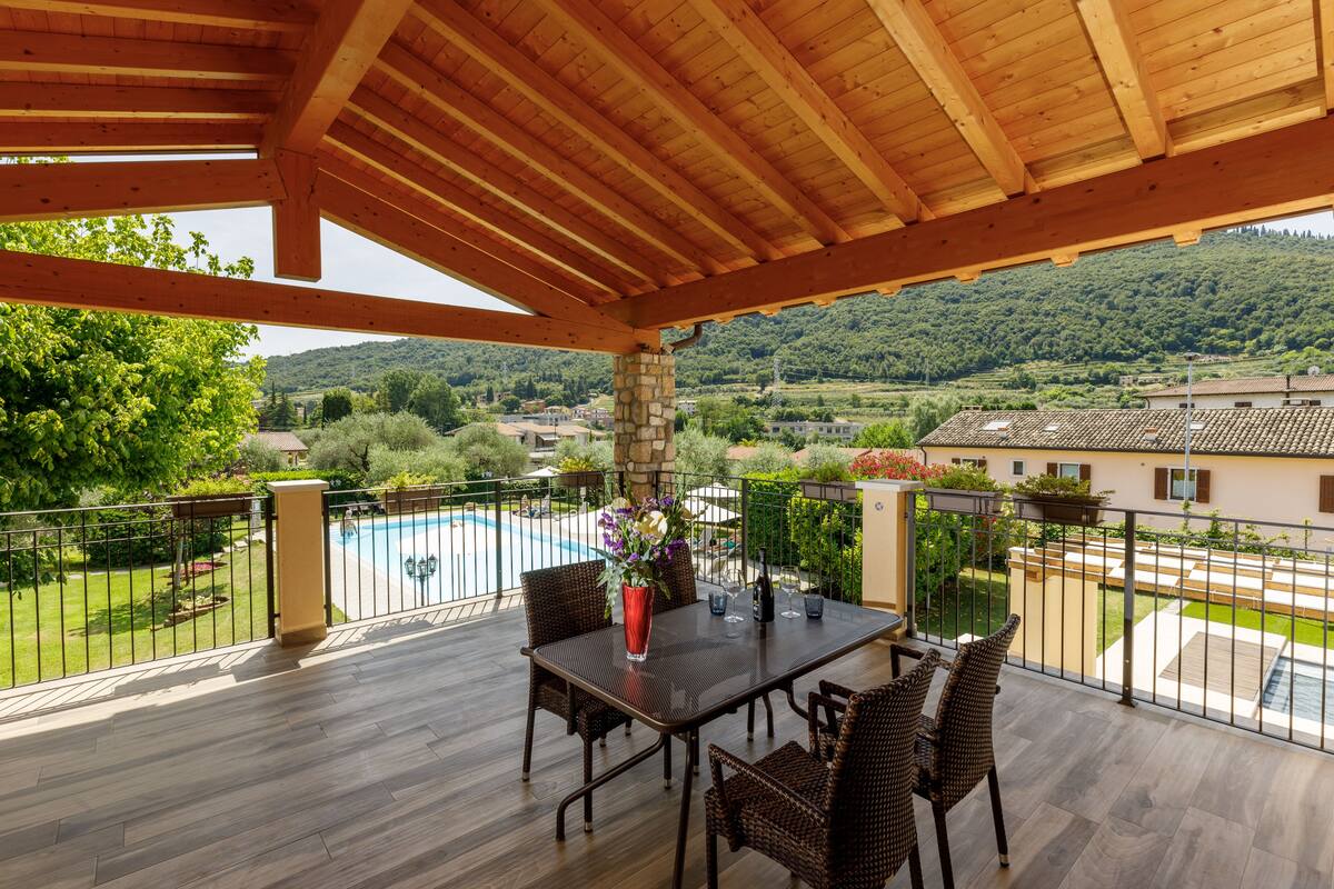 A covered outdoor area with a wooden ceiling, a table with chairs, and a view of a swimming pool and greenery.