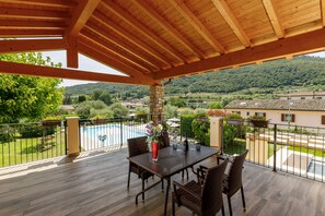 Un'area esterna coperta con tetto in legno, un tavolo con sedie e vista su una piscina e vegetazione.