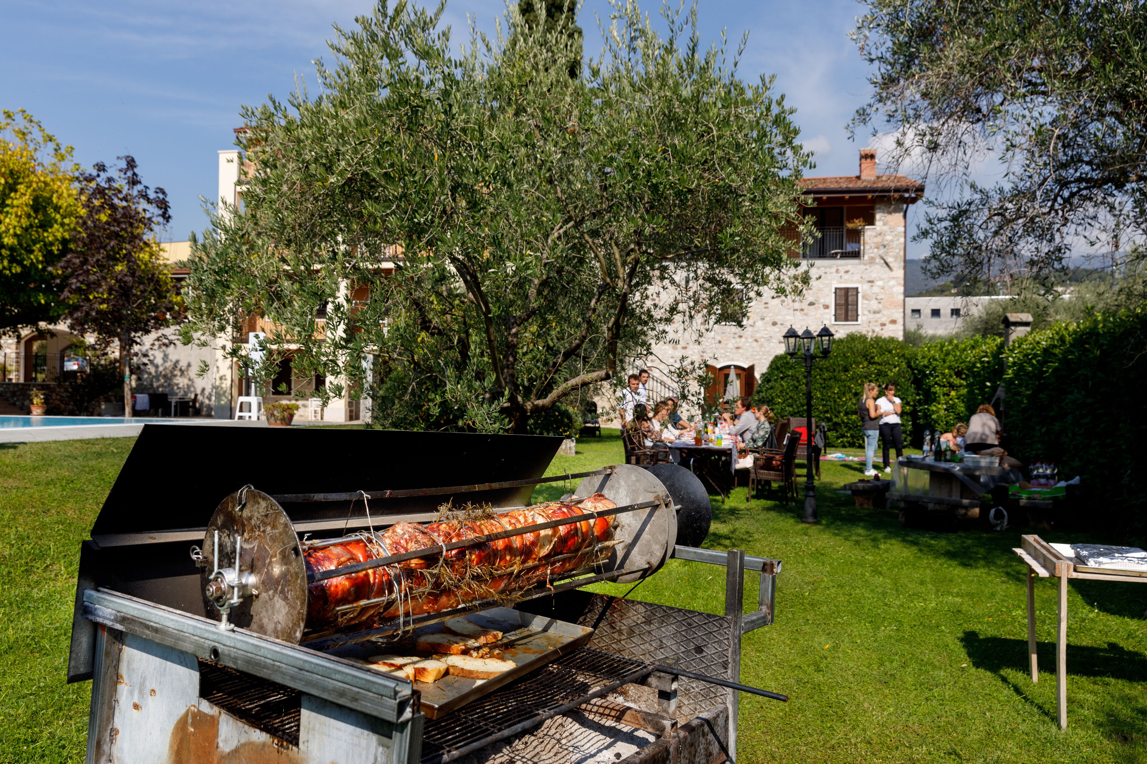Un grand barbecue extérieur avec une grande pièce de viande en train de cuire, entouré d’une zone herbeuse avec des arbres et un bâtiment en arrière-plan.