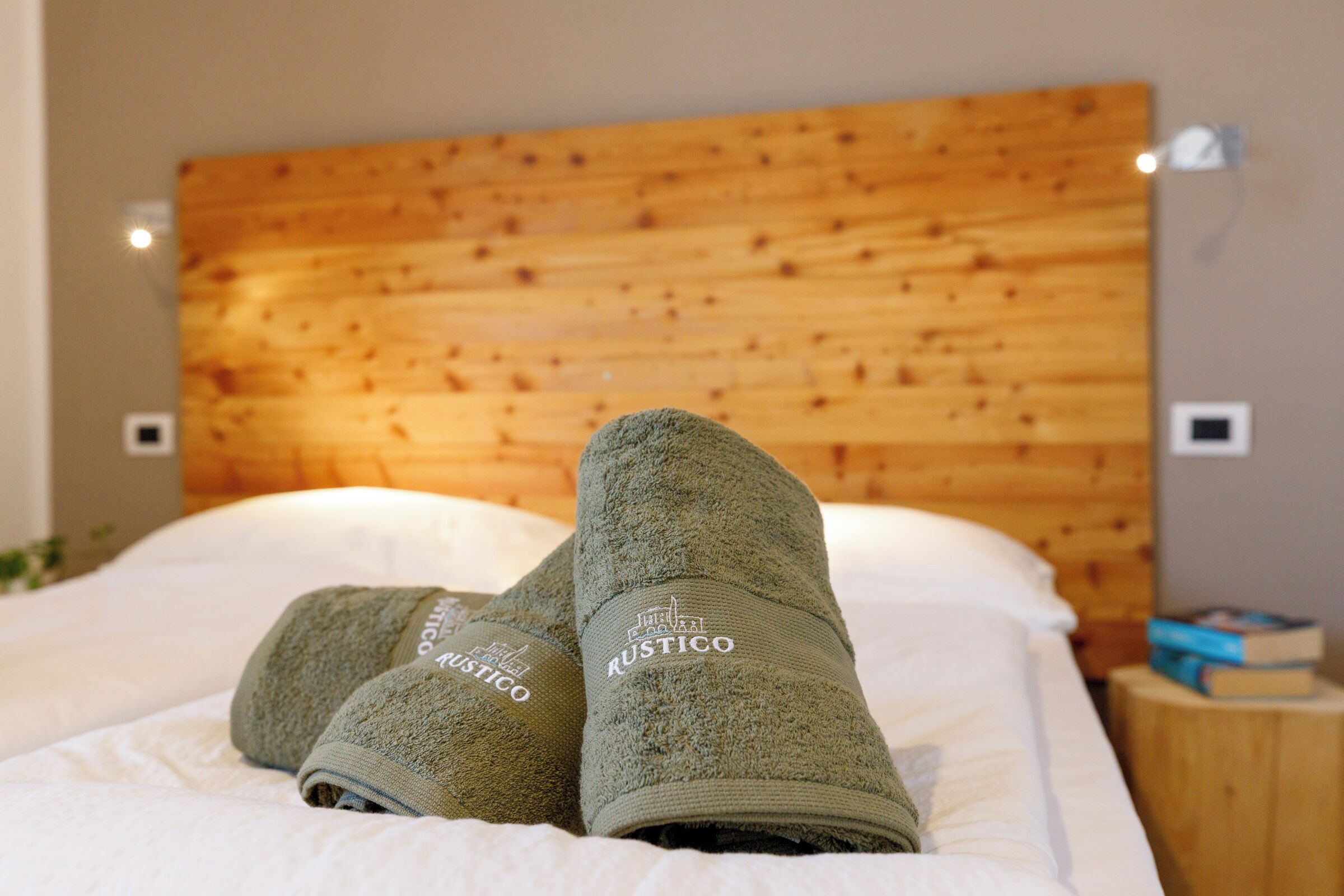 A bed with a wooden headboard and two rolled towels with the brand name "Rustico" on them.