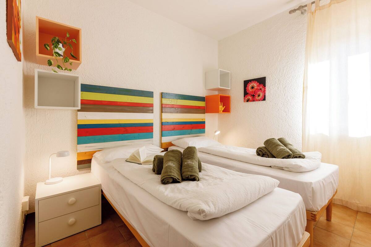 A hotel room with two beds, a nightstand, a bedside table, a lamp, a plant, and a colorful artwork on the wall.