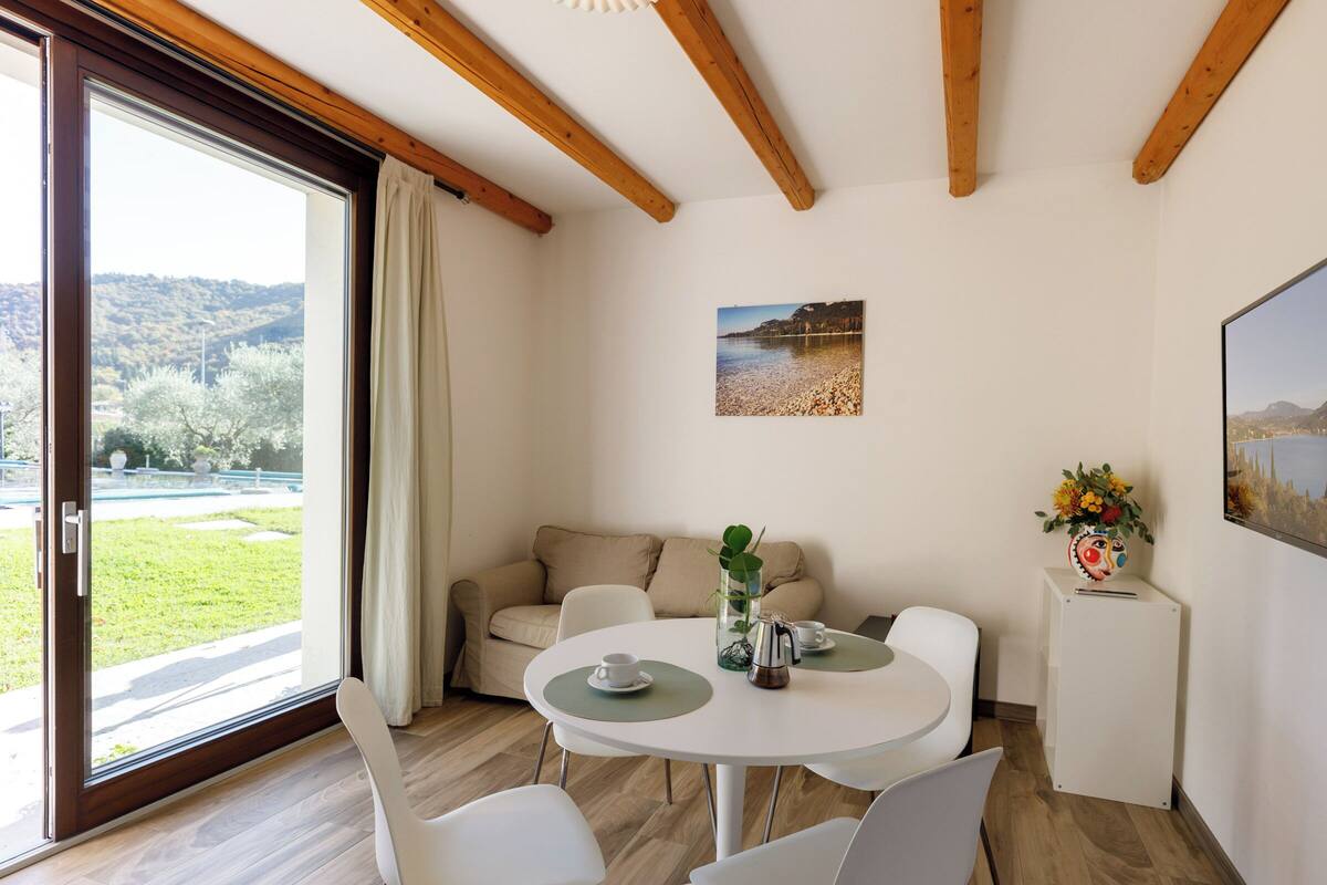 A modern room with a wooden ceiling, a round dining table with chairs, a sofa, and a large window with a view of greenery and hills.