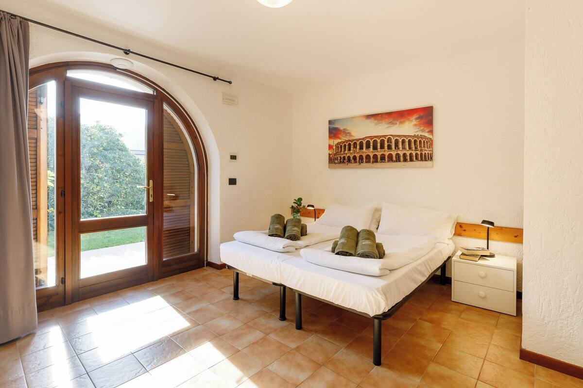 A bedroom with a bed, a nightstand, a painting of the Colosseum, and a sliding glass door to the outside.