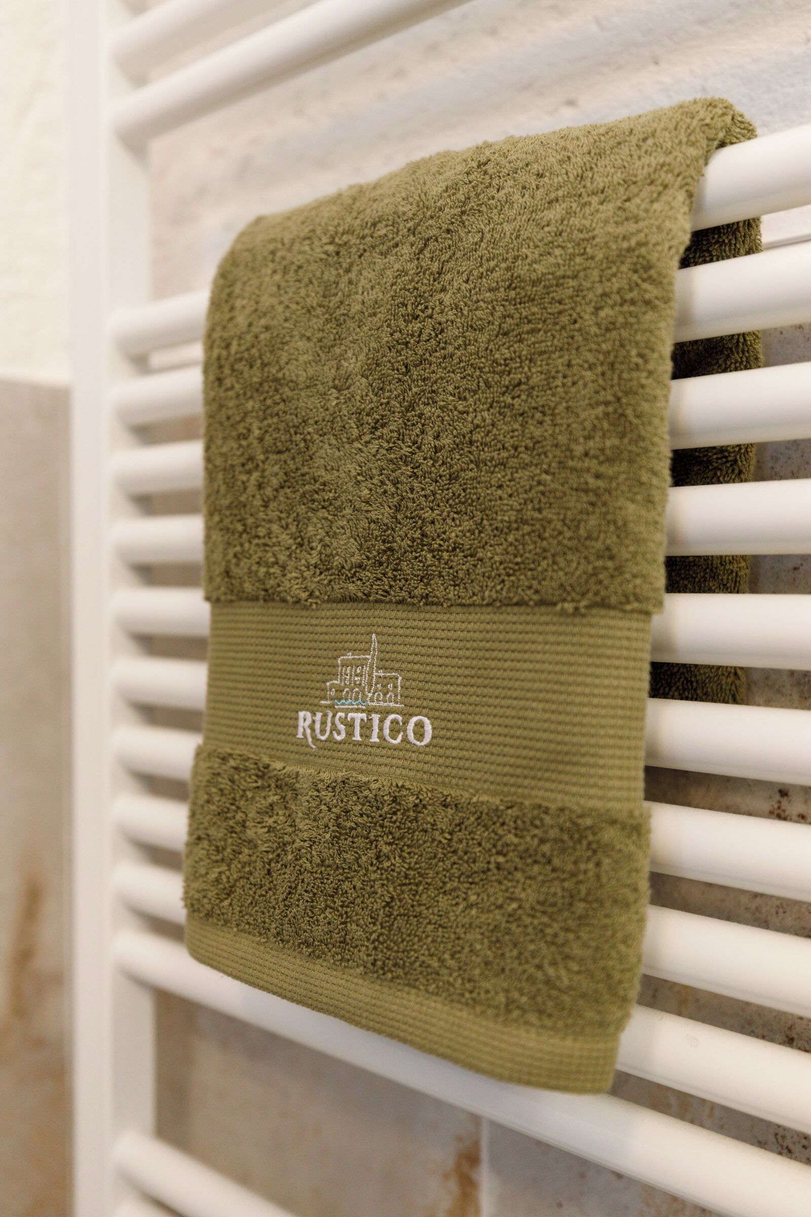 Un asciugamano verde piegato con un'etichetta che riporta la scritta "RUSTICO" appeso a un termosifone bianco.