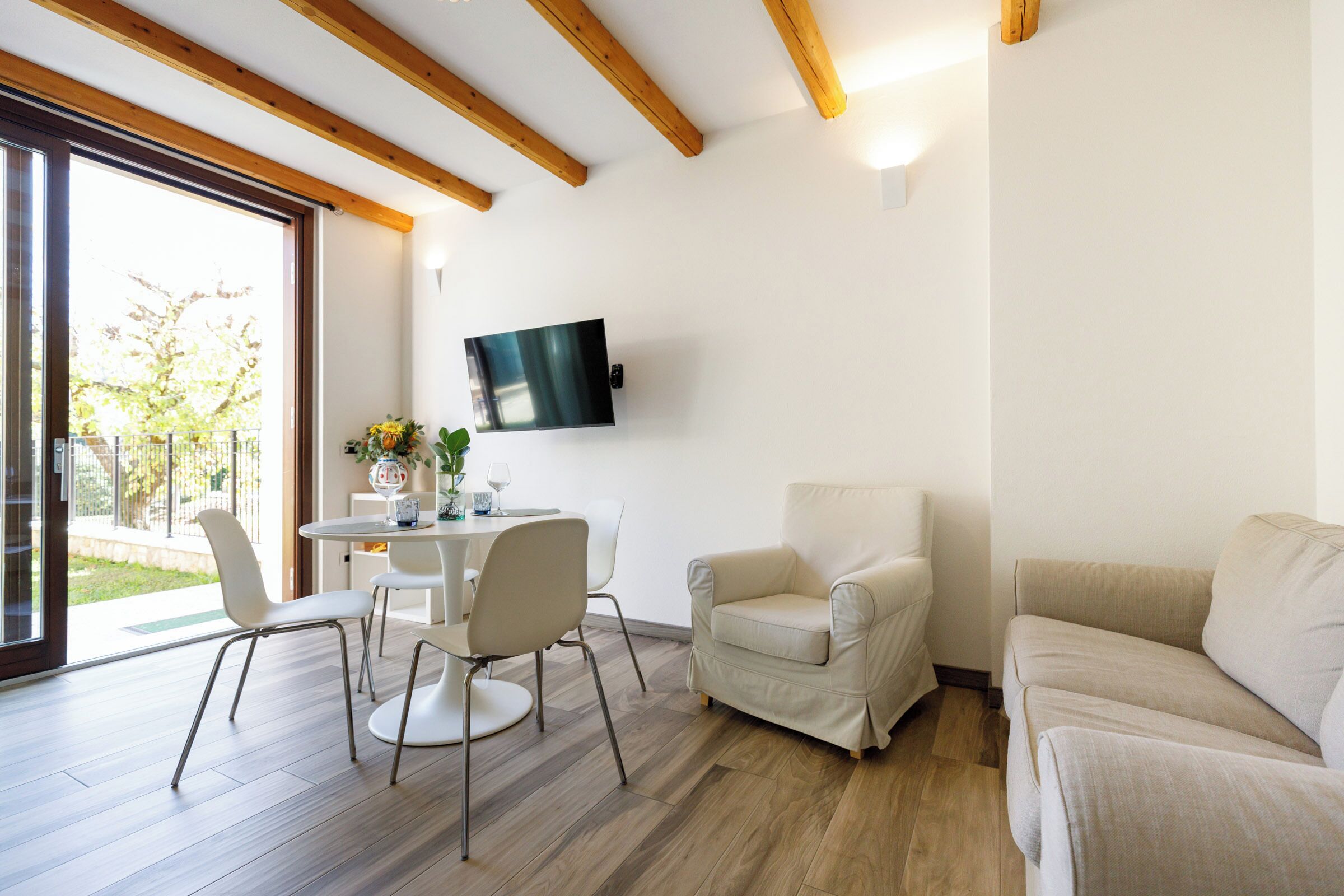 Una sala de estar moderna con techo de madera, un televisor de pantalla plana montado en la pared, una mesa de comedor redonda con sillas, un sofá color beige y un amplio ventanal corredera que da a un espacio exterior.