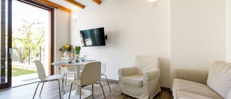 Una sala de estar moderna con techo de madera, un televisor de pantalla plana montado en la pared, una mesa de comedor redonda con sillas, un sofá color beige y un amplio ventanal corredera que da a un espacio exterior.