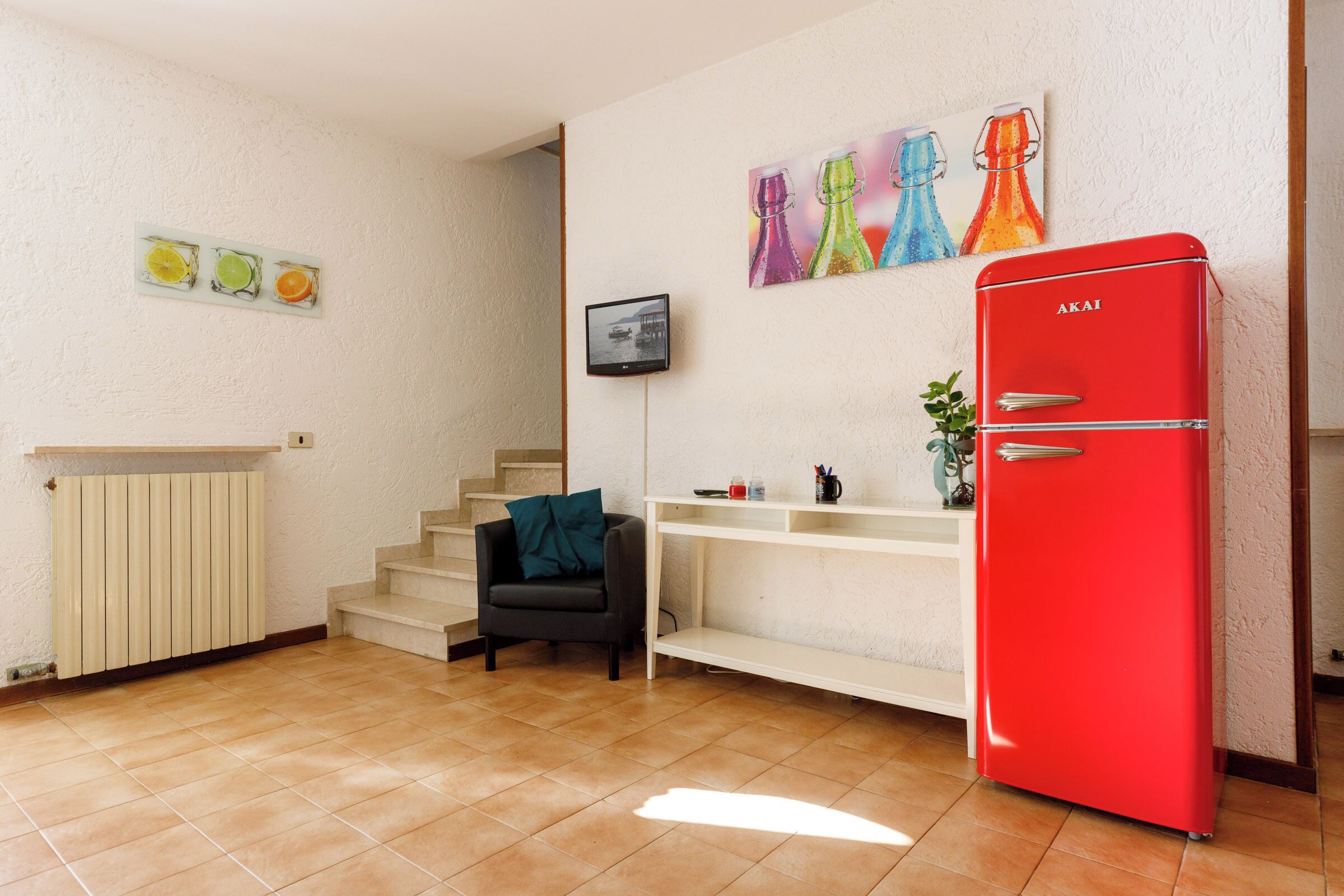 Un espace de vie compact comprenant un réfrigérateur rouge, un petit coin cuisine, un escalier et un coin salon avec une chaise et une plante.