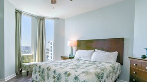 Hotelkamer met een groot bed, nachtkastje, lamp, stoel en uitzicht op de stad door grote ramen.