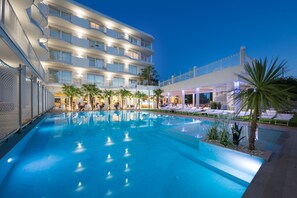 Area piscina di un hotel con una grande vasca, lettini prendisole e un edificio a più piani con balconi.