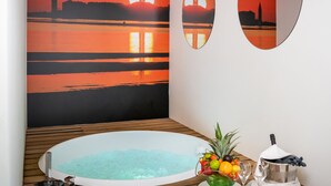 Une pièce avec une grande fresque murale représentant un coucher de soleil sur l’eau, un plancher en bois, un jacuzzi, un panier de fruits et deux miroirs.