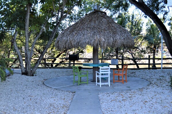 Relax in a serene, setting surrounded by trees and a fence, with a shaded thatched-roof structure and comfortable seating for two.