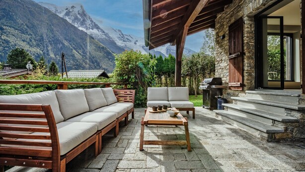 A patio with a wooden bench and a sofa, a table with cups and a bowl, and a grill.