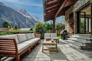 Une terrasse avec un banc en bois et un canapé, une table avec des tasses et un saladier, et un barbecue.