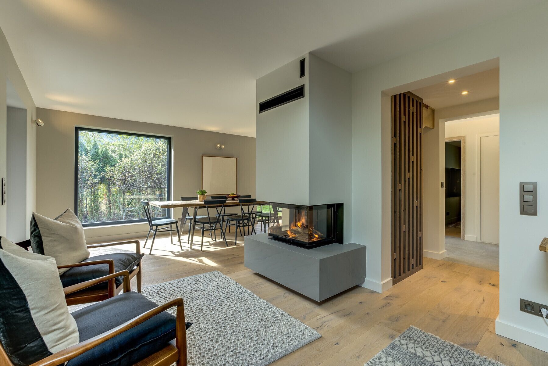 Een moderne woonkamer met een open haard, een eethoek en uitzicht op een bosrijk gebied door grote ramen.