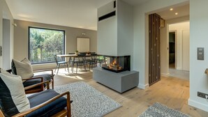 A modern living room with a fireplace, a dining area, and a view of a forest through large windows.