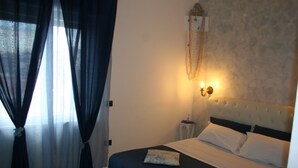 Un lit bien fait, avec une literie blanche et bleue, un luminaire fixé au mur et des rideaux légers à une fenêtre.