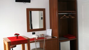 Une pièce de taille modeste, comprenant un bureau en bois, une chaise, une petite table recouverte d’une nappe rouge, un miroir, une armoire avec une tringle pour suspendre les vêtements et un petit réfrigérateur.