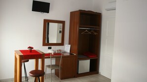 Une pièce de taille modeste, comprenant un bureau en bois, une chaise, un petit meuble de rangement, un miroir, un téléviseur et une armoire avec une tringle pour suspendre les vêtements.