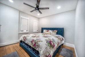 Une chambre à coucher avec un lit, un ventilateur de plafond et deux fenêtres équipées de stores.