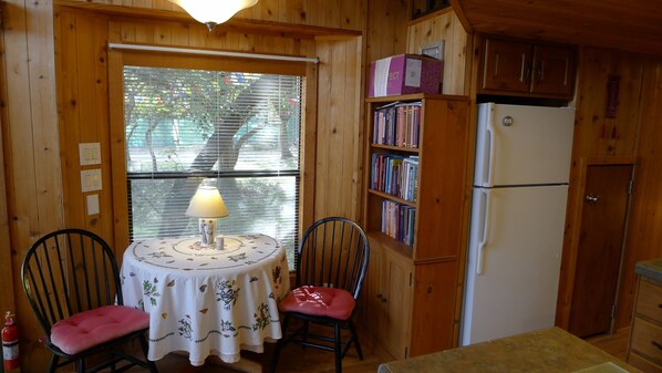 A wooden cabin interior with a dining table, chairs, a lamp, a bookshelf, and a refrigerator.